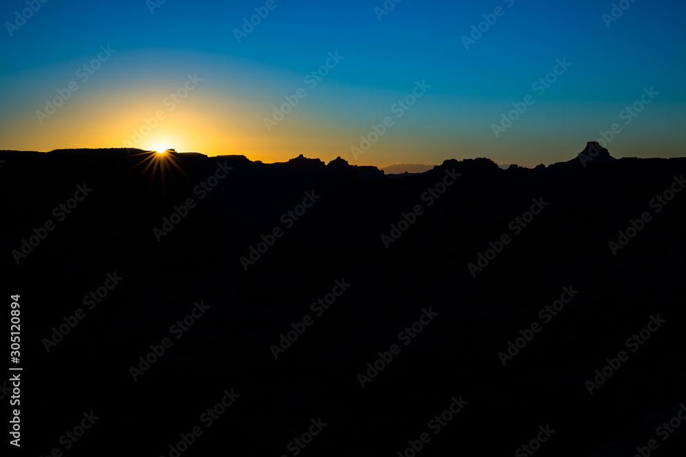 Sun rising over black foreground with jagged desert plateau and mesas silhouetted against the blue sky.