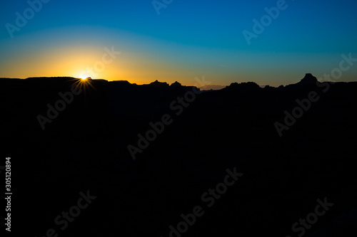 Sun rising over black foreground with jagged desert plateau and mesas silhouetted against the blue sky. photo