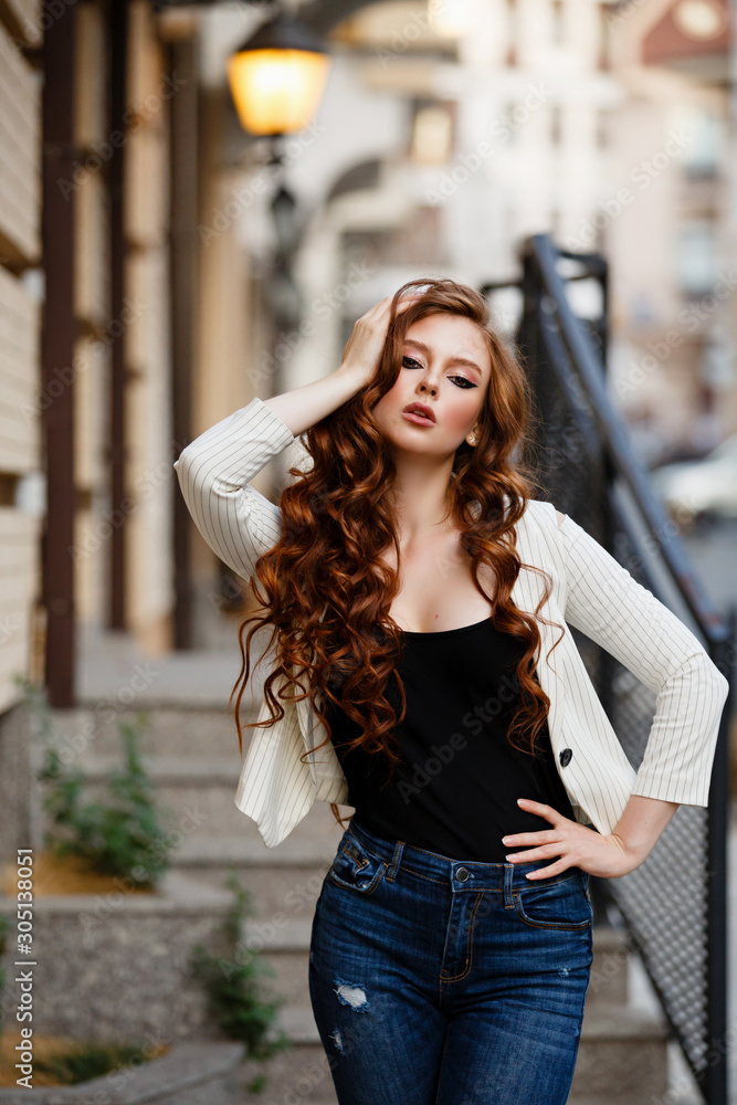 Young curled girl weared in white jacket and jeans on background of old City. Fashion style girl with long curled red hair. Fashion walk in the City. Street style Modern strict urban girl on stairs