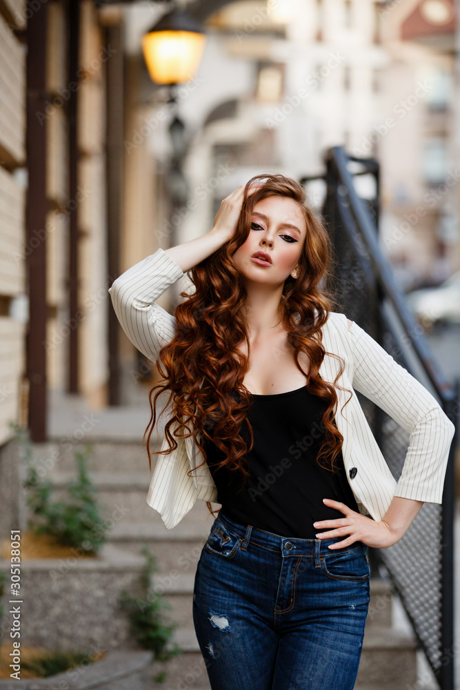 Young curled girl weared in white jacket and jeans on background of old City. Fashion style girl with long curled red hair. Fashion walk in the City. Street style Modern strict urban girl on stairs Young curled girl weared in white jacket and jeans on background of old City. Fashion style girl with long curled red hair. Fashion walk in the City. Street style Modern strict urban girl on stairs