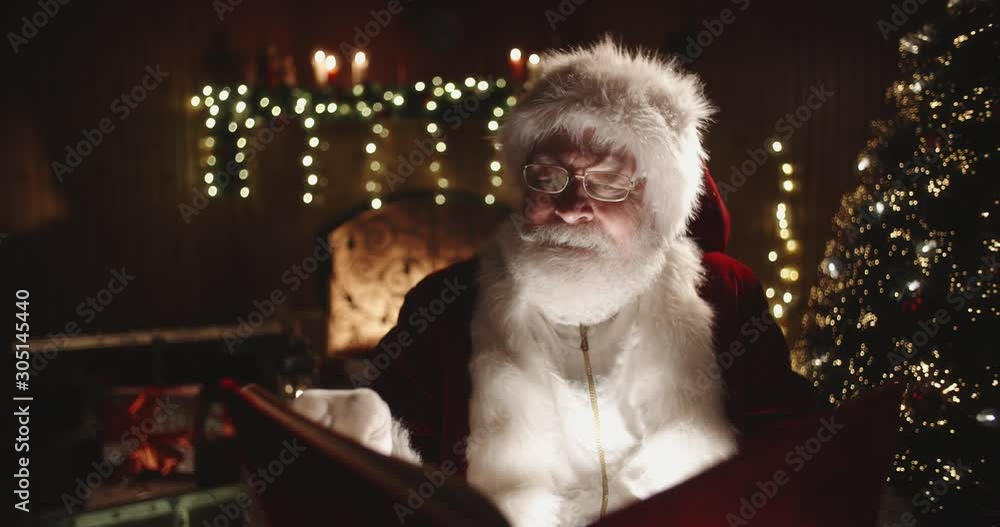 Joyful authentic santa clause flipping through pages of red covered book, with fireplace and christmas tree on background - christmas spirit concept close up 4k footage Joyful authentic santa clause flipping through pages of red covered book, with fireplace and christmas tree on background - christmas spirit concept close up 4k footage