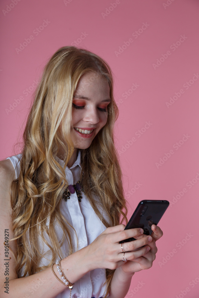 curly young blonde girl with long hair holding smartphone and writing in chat, Studio with pink background curly young blonde girl with long hair holding smartphone and writing in chat, Studio with pink background