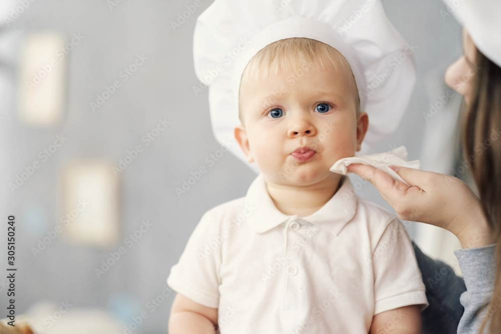 mother and child on kitchen, white hats of chef, mom wipes baby with help of napkin mother and child on kitchen, white hats of chef, mom wipes baby with help of napkin