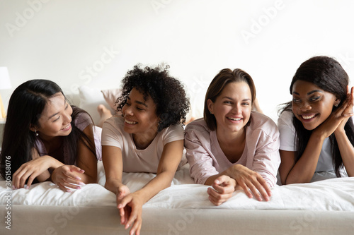 Happy diverse young ladies relax on bed enjoy pajama party