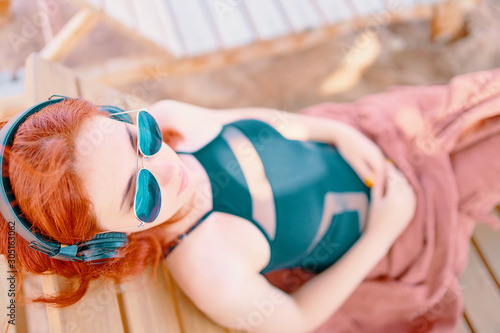 Cute woman listening to music on the beach. Redhead girl listening melody with headphones and smartphone while relaxing on lounger near ocean.