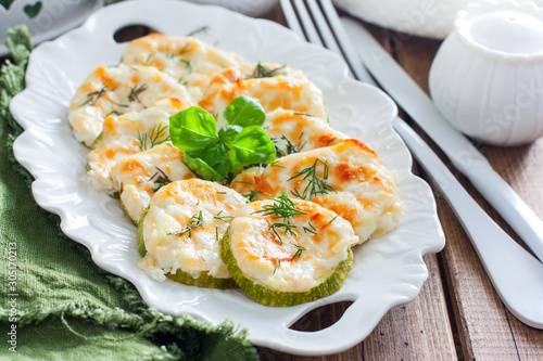 Baked zucchini with cheese slices on a white dish on a wooden table, horizontal