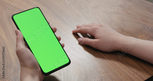 Young woman sitting at a table and using a smartphone with vertical green screen