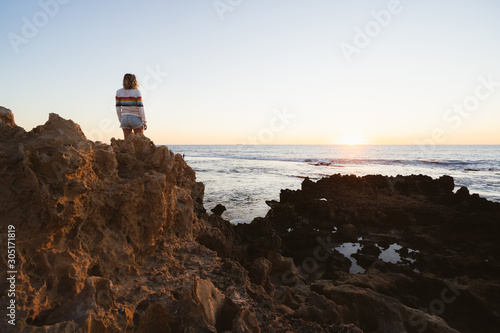 Young blonde woman standing on jagged rocks, wearing a rainbow jumper, watching the sunset over the beautiful beach and ocean at Trigg beach, Perth, Western Australia. 