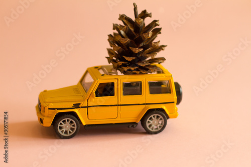 Yellow toy car with a forest cone on the roof on a soft pink background with Christmas toys. postcard, gift delivery. A gift for Christmas and new year. space for text.