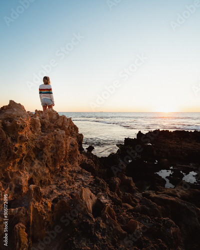 Young blonde woman standing on jagged rocks, wearing a rainbow jumper, watching the sunset over the beautiful beach and ocean at Trigg beach, Perth, Western Australia. 