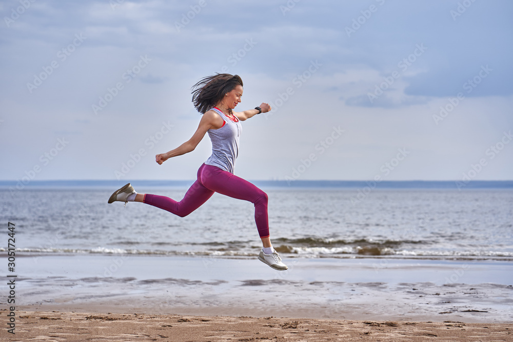 Young woman with black hair jumps while jogging. A woman is engaged in gymnastics in the spring morning on the sandy bank of a large river. Cloudy morning. Young woman with black hair jumps while jogging. A woman is engaged in gymnastics in the spring morning on the sandy bank of a large river. Cloudy morning.
