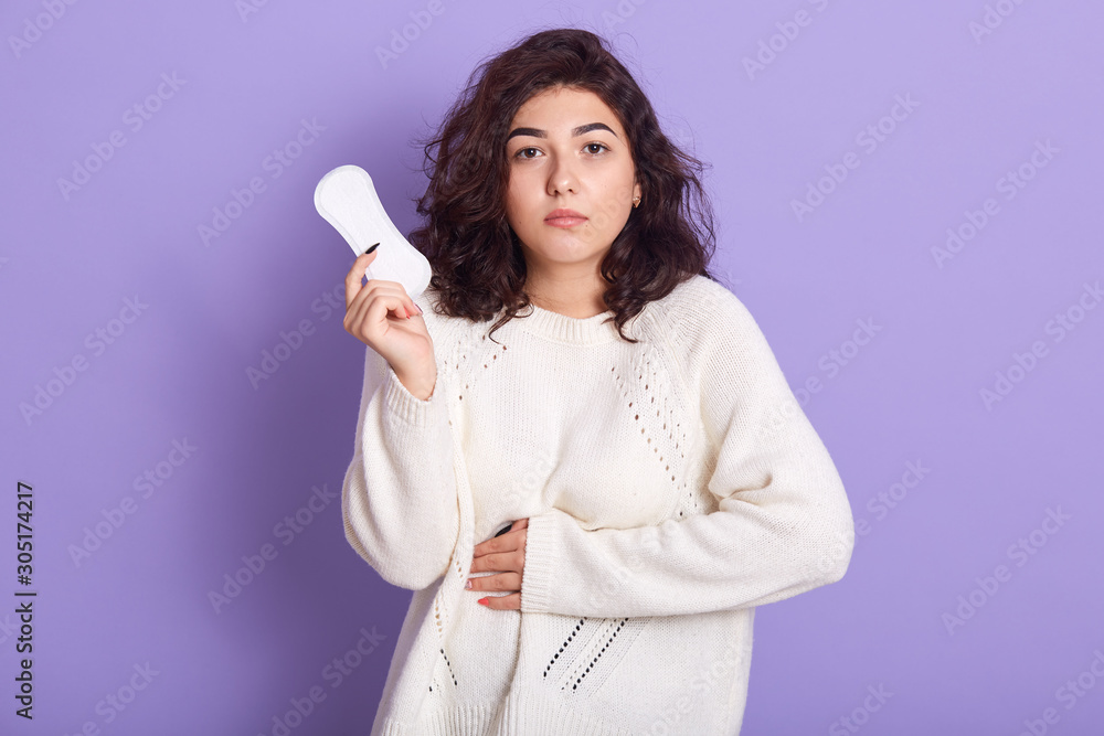 Gloomy Caucasian woman cares about personal hygiene, holds sanitary pad in hands, suffers from pain during menstruation, has sad expression, wear white outfit, poses in studio isolated over lilac wall Gloomy Caucasian woman cares about personal hygiene, holds sanitary pad in hands, suffers from pain during menstruation, has sad expression, wear white outfit, poses in studio isolated over lilac wall