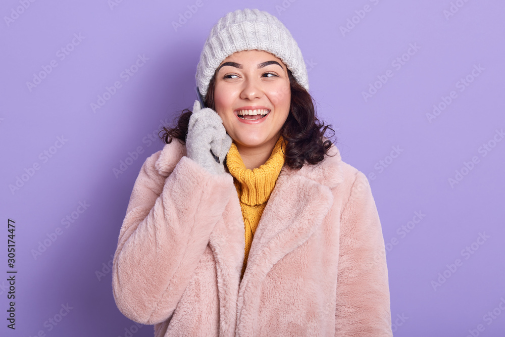 Picture of good looking beautiful model with dark curly hair looking aside, smiling sincerely, keeping smartphone in one hand, wearing pink faux fur coat, yellow sweater, light gloves and hat. Picture of good looking beautiful model with dark curly hair looking aside, smiling sincerely, keeping smartphone in one hand, wearing pink faux fur coat, yellow sweater, light gloves and hat.