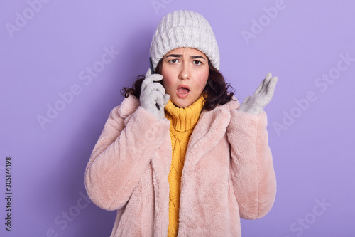 Shocked outraged young woman standing isolated over lilac background in studio, gossiping over mobile phone, putting her smartphone close to ear, opening her mouth widely. Breaking news concept. photo