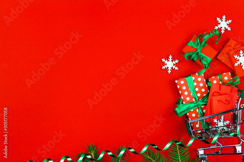 Small trolley, mini shopping cart full of presents. Gift boxes in wrapping paper with green ribbons, packages, christmas tree branch on red background. New year fair, market, sale concept.