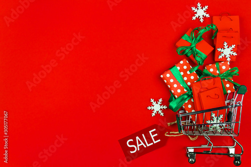 Small trolley  mini shopping cart full of Christmas presents and packages. Gift boxes are packed in decorative paper and green ribbons on red background. New year fair  market  sale concept.