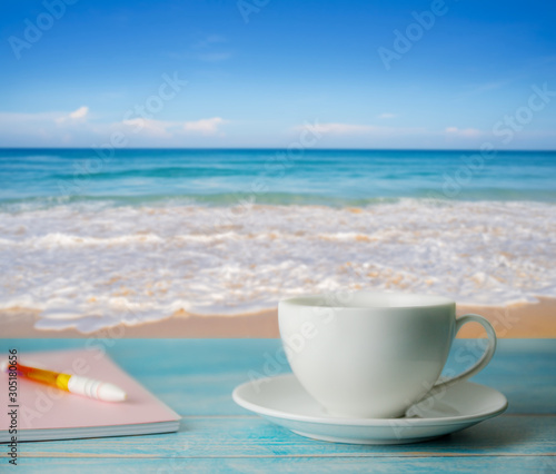 A cup of coffee and notebook with blurred beach sand and blue sky