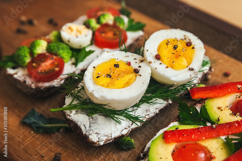 three bruschettas on dark bread with curd cheese, eggs, vegetables, spices and herbs on a wooden board