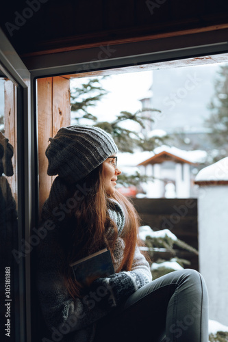 Beautiful woman in a sweater sitting home by the window. Blurred winter snow tree background. Morning, coziness, winter and people concept