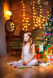 Cute little girl sits on the floor in the evening in the room near the festive Christmas tree and dreams. Christmas and New Year.