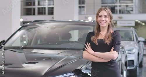 Attractive Caucasian woman with pleasant smile posing next to the new black automobile. Nice girl buying or renting car in car dealership. Car rental, car business. Cinema 4k footage ProRes HQ.