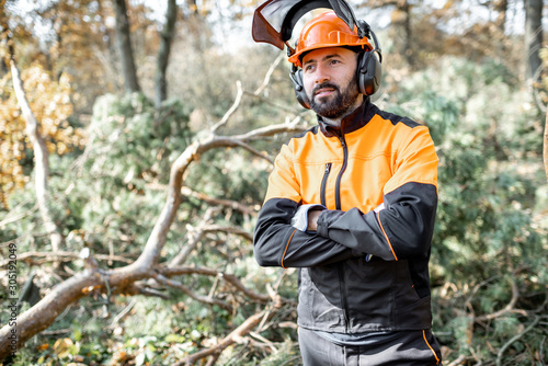 Waist-up portrait of a professional lumberman in harhat and protective workwear standing in the pine forest