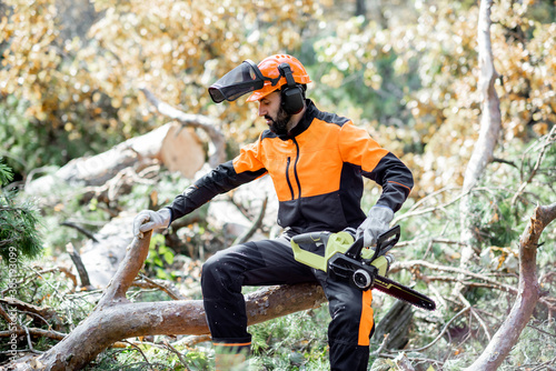 Portrait of a professional lumberman in protective workwear sitting with a chainsaw on the felled tree, resting after the hard work in the forest