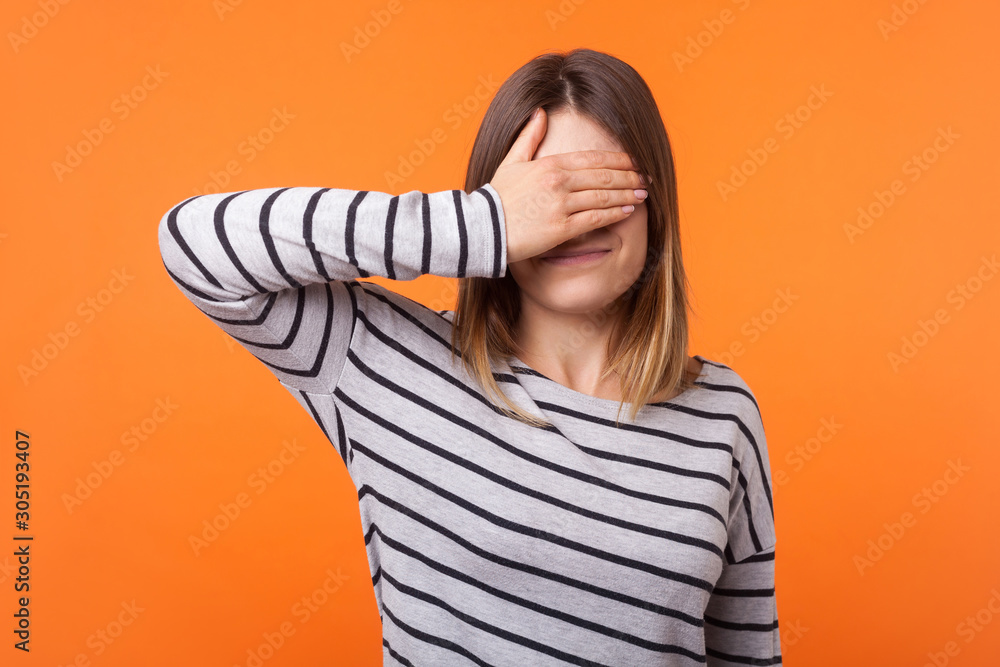 Don't want to look at this. Portrait of young scared woman with brown hair in long sleeve shirt standing, covering eyes with hand, refusing to watch. indoor studio shot isolated on orange background Don't want to look at this. Portrait of young scared woman with brown hair in long sleeve shirt standing, covering eyes with hand, refusing to watch. indoor studio shot isolated on orange background