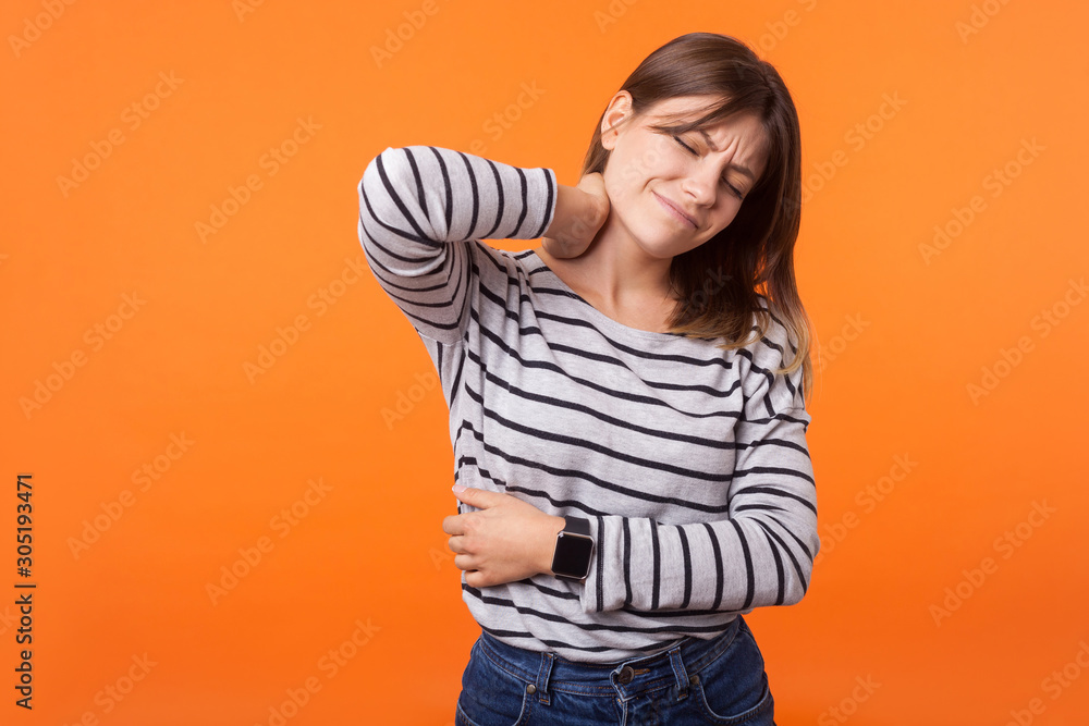 Portrait of tired sick woman with brown hair in long sleeve striped shirt standing with closed eyes, rubbing neck to relieve pain, muscle strain. indoor studio shot isolated on orange background Portrait of tired sick woman with brown hair in long sleeve striped shirt standing with closed eyes, rubbing neck to relieve pain, muscle strain. indoor studio shot isolated on orange background