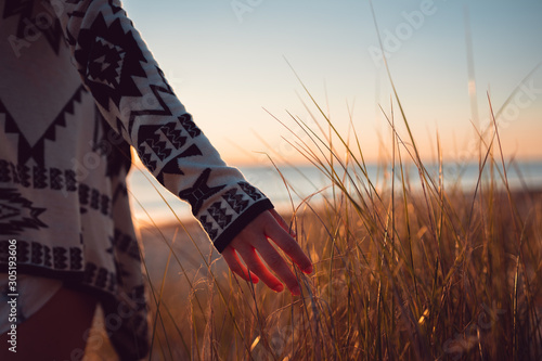 Young woman wearing a jersey with her hand exploring the sand dune grass on the beach with the sunset in the background over the ocean, Cottesloe, Perth, Western Australia.
