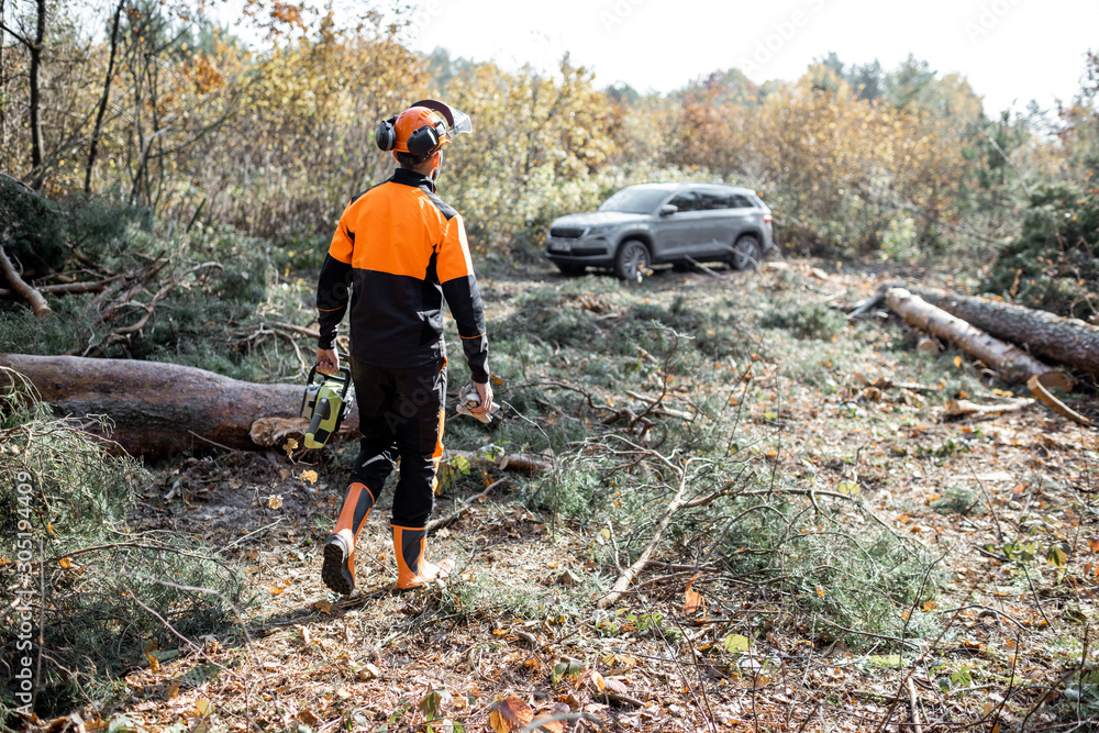 Professional lumberman in protective clothes making woods logging with chainsaw, going to his SUV car in the forest Professional lumberman in protective clothes making woods logging with chainsaw, going to his SUV car in the forest