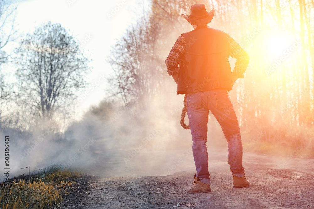 Fototapeta premium A man is wearing a cowboy hat and a loso in the field. American farmer in a field wearing a jeans hat and with a lasso in the smoke of a fire. A man walks through a burning field