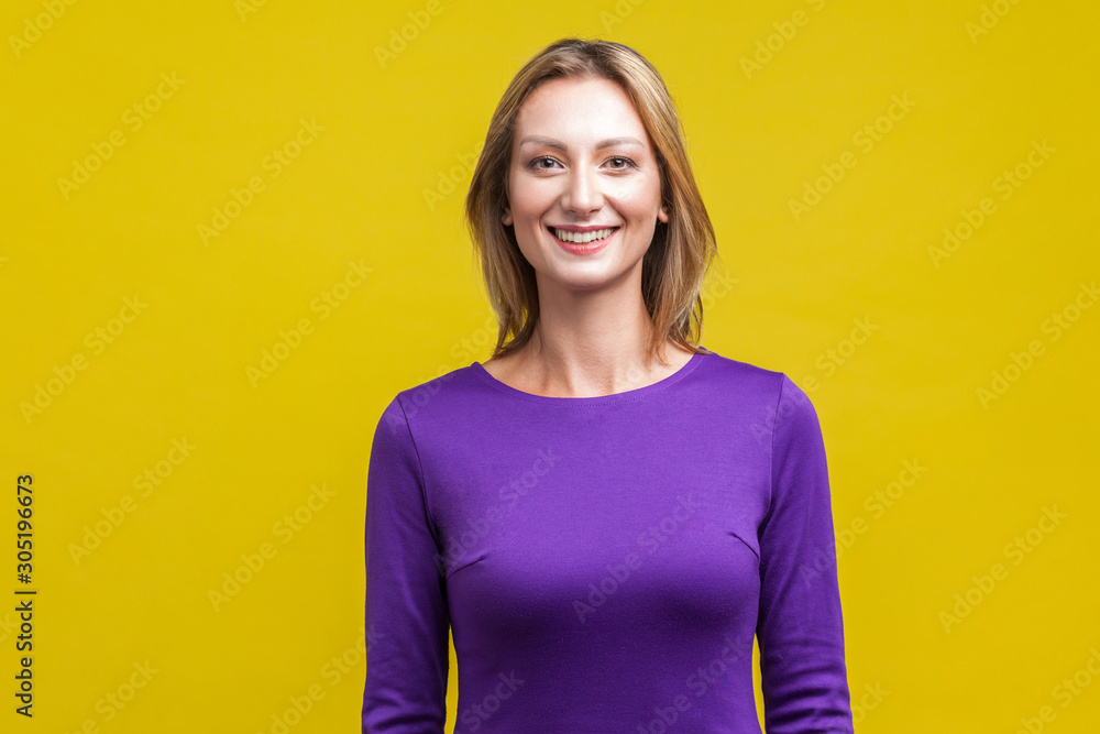 Fototapeta premium Portrait of positive elegant woman in tight purple dress standing, looking at camera with cute engaging toothy smile, beautiful with clean shiny skin. indoor studio shot isolated on yellow background