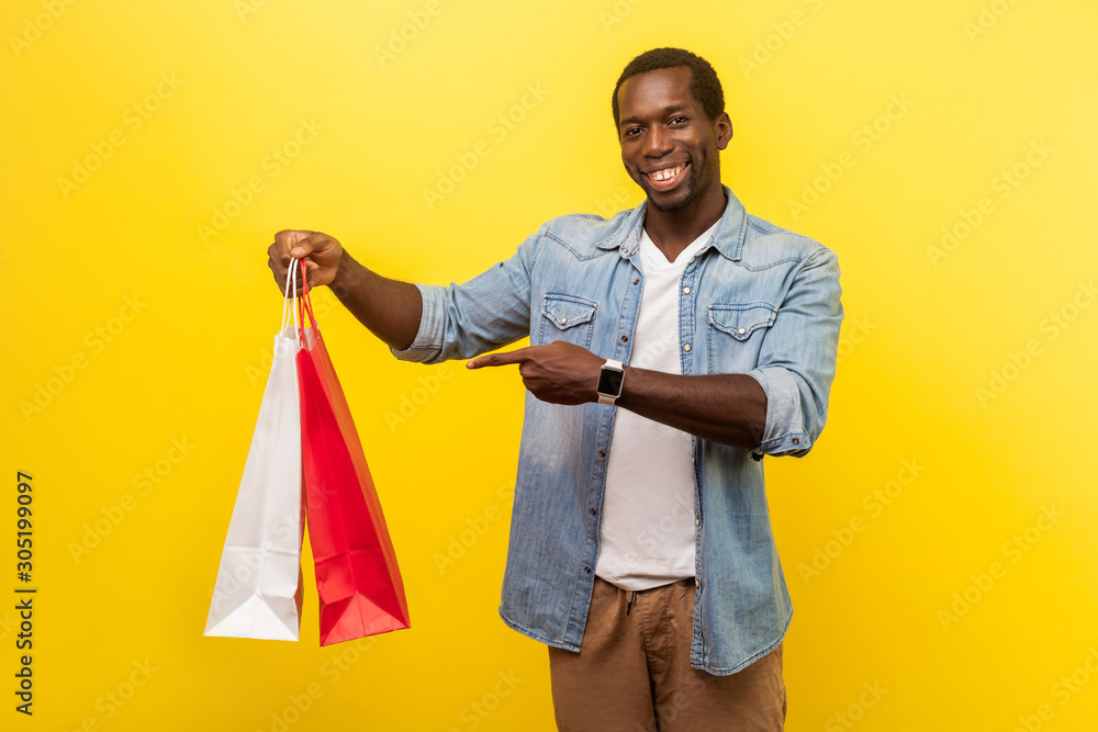 Portrait of happy satisfied male buyer in denim casual shirt with rolled up sleeves pointing at shopping bags and looking at camera with toothy smile. indoor studio shot isolated on yellow background Portrait of happy satisfied male buyer in denim casual shirt with rolled up sleeves pointing at shopping bags and looking at camera with toothy smile. indoor studio shot isolated on yellow background