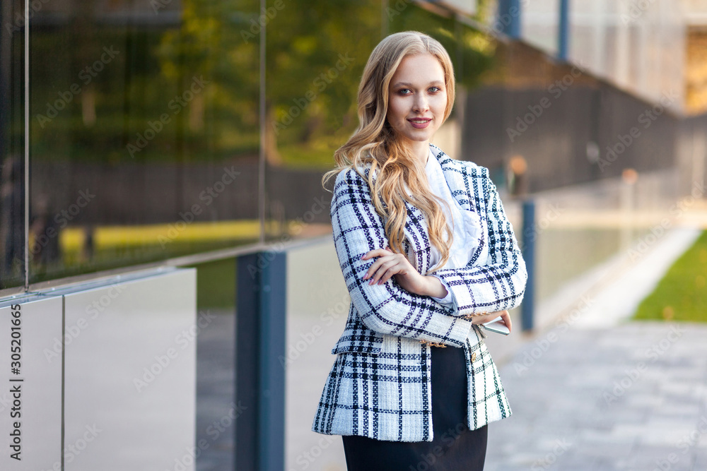 Portrait of attractive confident businesslady with long blond hair in plaid jacket and skirt standing with crossed arms and looking at camera with toothy smile. outdoors, near business center building Portrait of attractive confident businesslady with long blond hair in plaid jacket and skirt standing with crossed arms and looking at camera with toothy smile. outdoors, near business center building