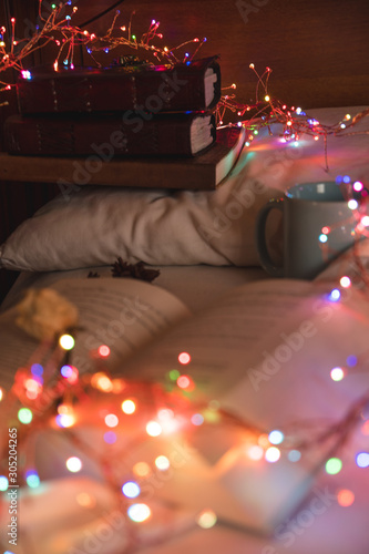 Cozy background of an open book on a bed, next to a mug and surrounded by colorful christmas lights