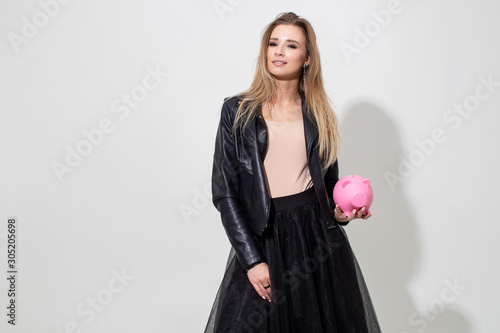 A girl in a black leather jacket holds a pink piggy bank on a white background