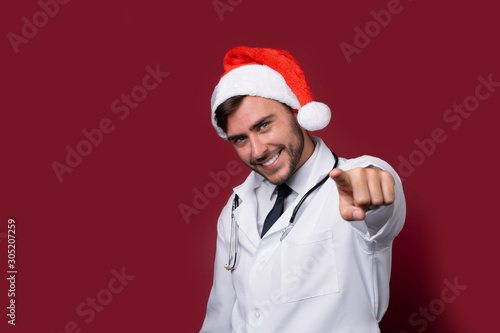 Young handsome doctor in white uniforme and Santa Claus hat standing in studio on red background smile and finger in camera
