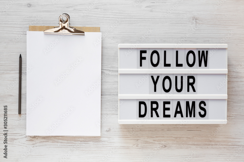 'Follow your dreams' words on a lightbox, clipboard with blank sheet of paper on a white wooden background, top view. Overhead, from above, flat lay. Copy space.