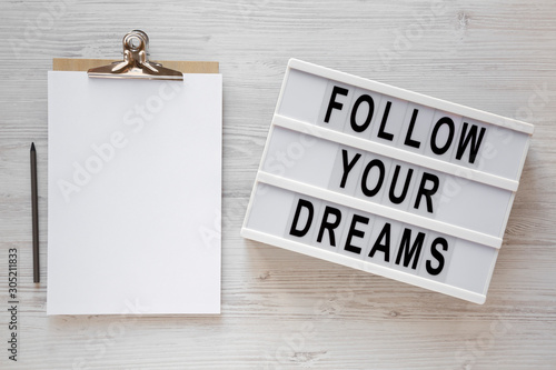 'Follow your dreams' words on a lightbox, clipboard with blank sheet of paper on a white wooden surface, top view. Overhead, from above, flat lay.