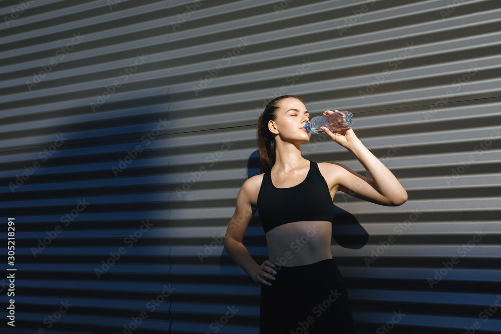 Healthy female standing outdoors and drinking water after exercising session. Fitness woman taking a break after workout