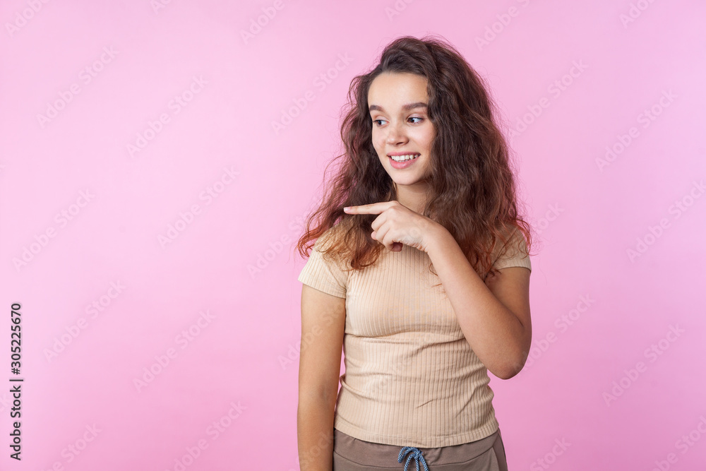 Portrait of happy beautiful teen girl with long curly brunette hair wearing casual style beige clothes pointing to the side and smiling, showing empty copy space. studio shot isolated, pink background Portrait of happy beautiful teen girl with long curly brunette hair wearing casual style beige clothes pointing to the side and smiling, showing empty copy space. studio shot isolated, pink background