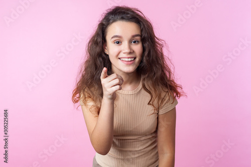 Hey you! Portrait of cheerful teenage girl with curly brunette hair wearing casual style beige clothes pointing at camera and smiling genuinely, choosing you. studio shot isolated on pink background