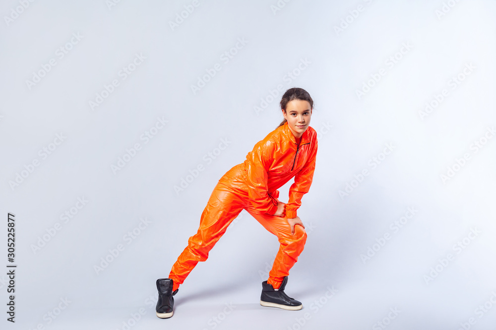 Full length image of energetic teenage girl with brunette hair wearing bright orange jumpsuit dancing, showing butterfly hip hop move, hobby activities. indoor studio shot isolated on white background