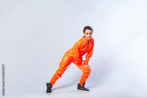 Full length image of energetic teenage girl with brunette hair wearing bright orange jumpsuit dancing, showing butterfly hip hop move, hobby activities. indoor studio shot isolated on white background © khosrork