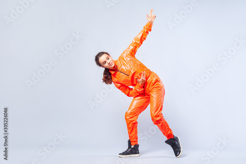 Fototapeta Naklejka Na Ścianę i Meble -  Full length portrait of positive energetic teen girl with brunette hair in bright orange jumpsuit dancing hip-hop and pointing up with finger pistol, hobby activities. studio shot, white background
