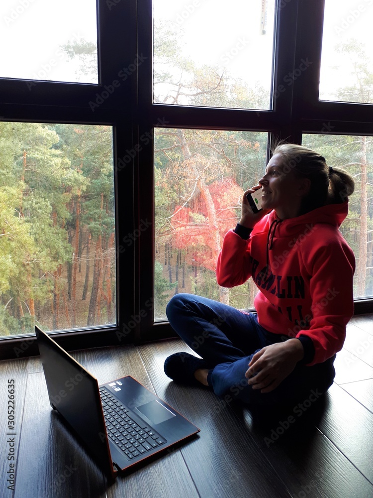 young fair-skinned girl at home working on a laptop and talking on the phone near a large window young fair-skinned girl at home working on a laptop and talking on the phone near a large window