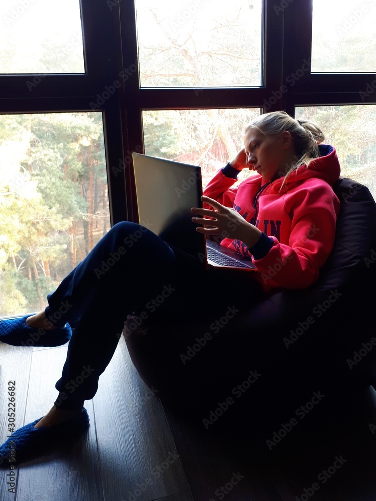 young fair-skinned girl at home working on a laptop and talking on the phone near a large window young fair-skinned girl at home working on a laptop and talking on the phone near a large window