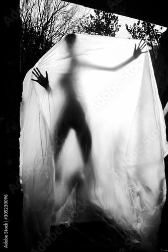 Slender nude girl, covered by a cellophane film, through which only her silhouette is visible and her nakedness is covered, in the ruins of a destroyed building. Conceptual, artistic, creative design.
