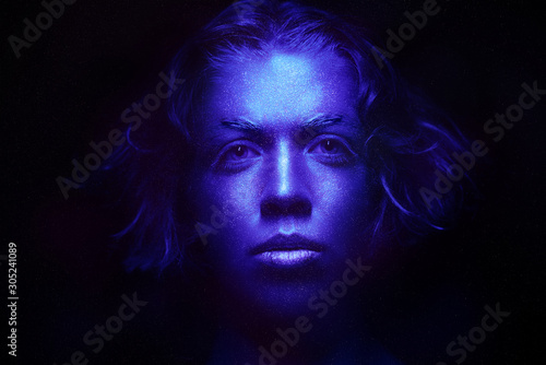 Beautiful girl in a shiny metallic space creative make-up of blue hue. Dark background, studio portrait of a model's face in blue paint. full-face portrait symmetrical with beautiful curly hair.